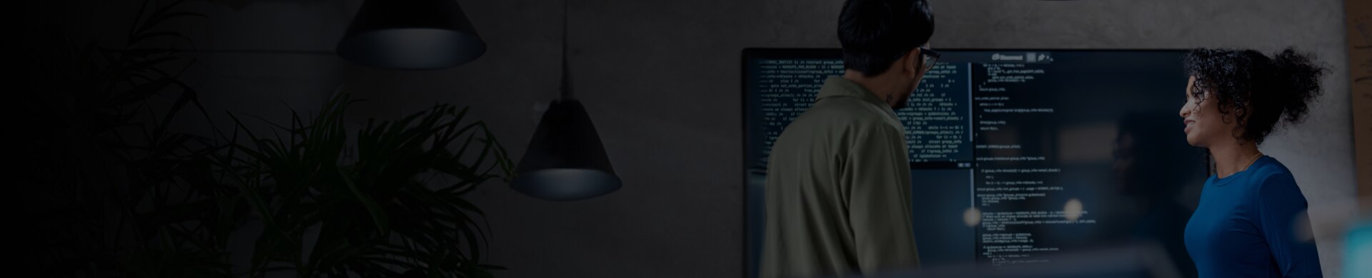 Two developers discussing code displayed on a large screen in a dim workspace.