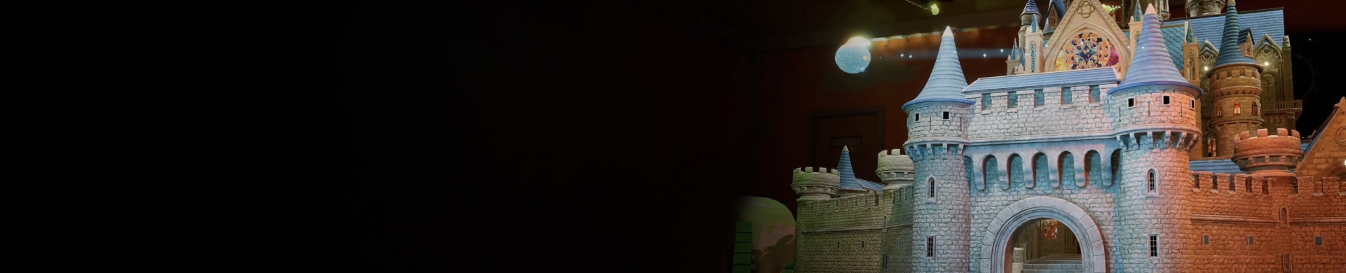 Illuminated fairy-tale castle at night with stone towers and an arched gate, fading into darkness on the left.